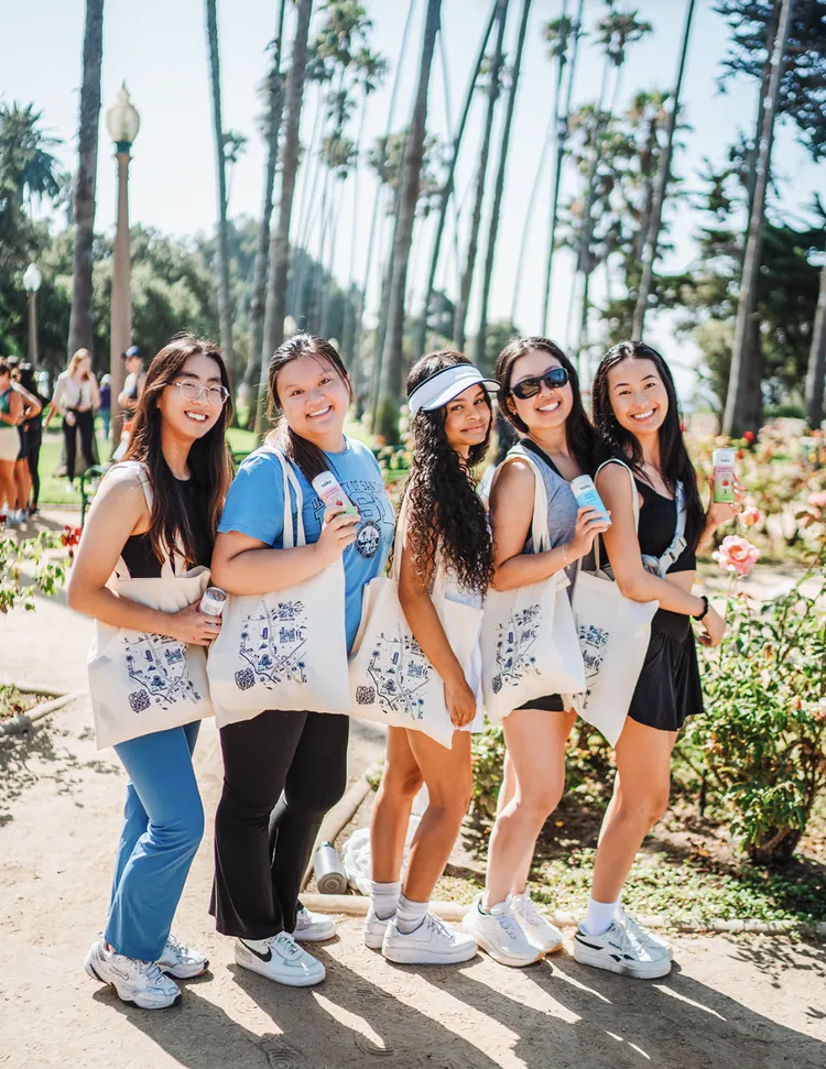 Group of women with tote bags at an outdoor event under palm trees